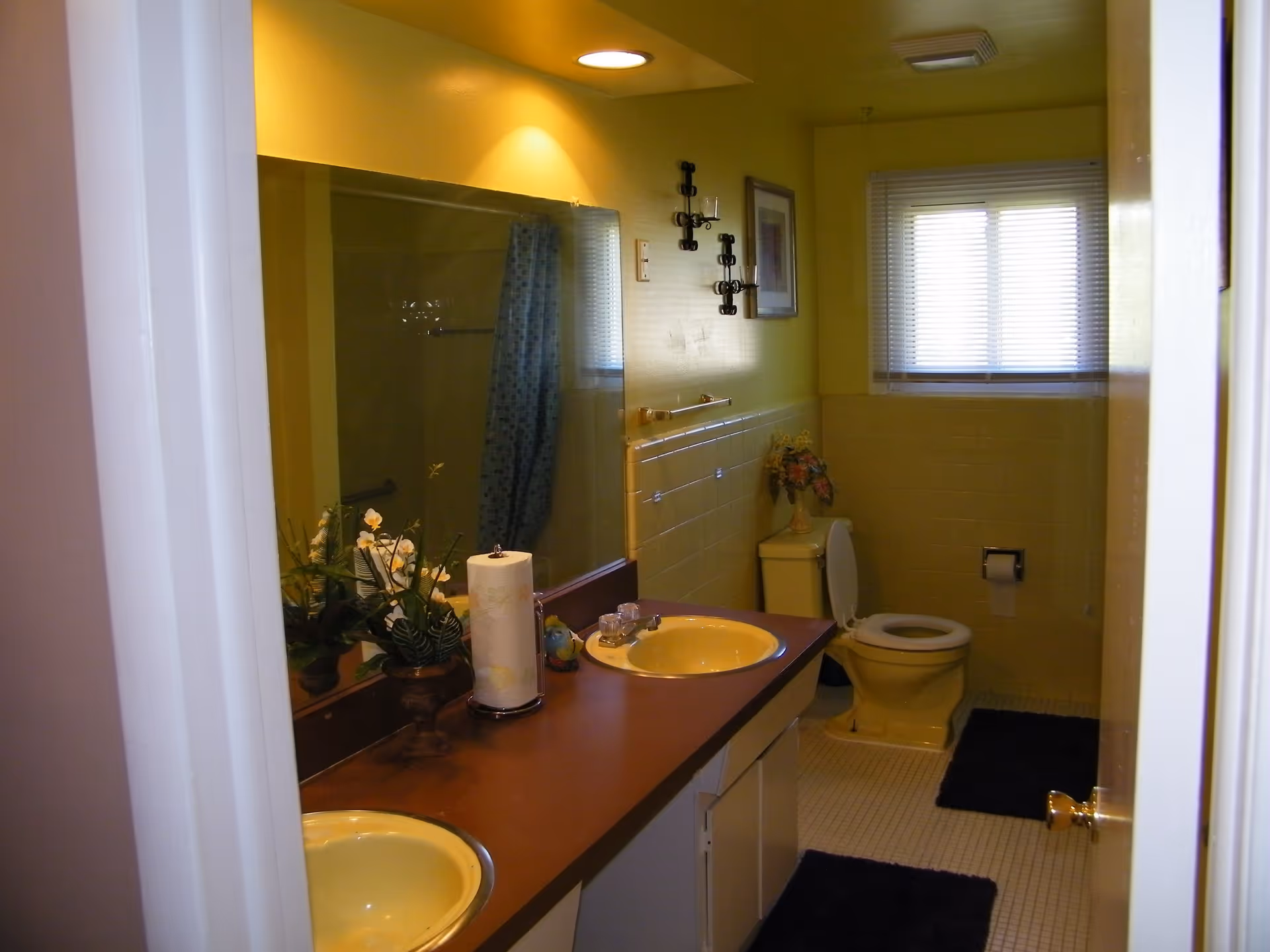 Interior view of a bathroom with yellow walls and fixtures, including two yellow sinks on a brown countertop, a toilet with the lid open, a large mirror above the sinks, a window with blinds, and decorative items such as a flower vase and wall-mounted candle holders.