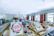Bright common activity room with tables arranged for painting, easels displaying floral canvases, art supplies and seating.