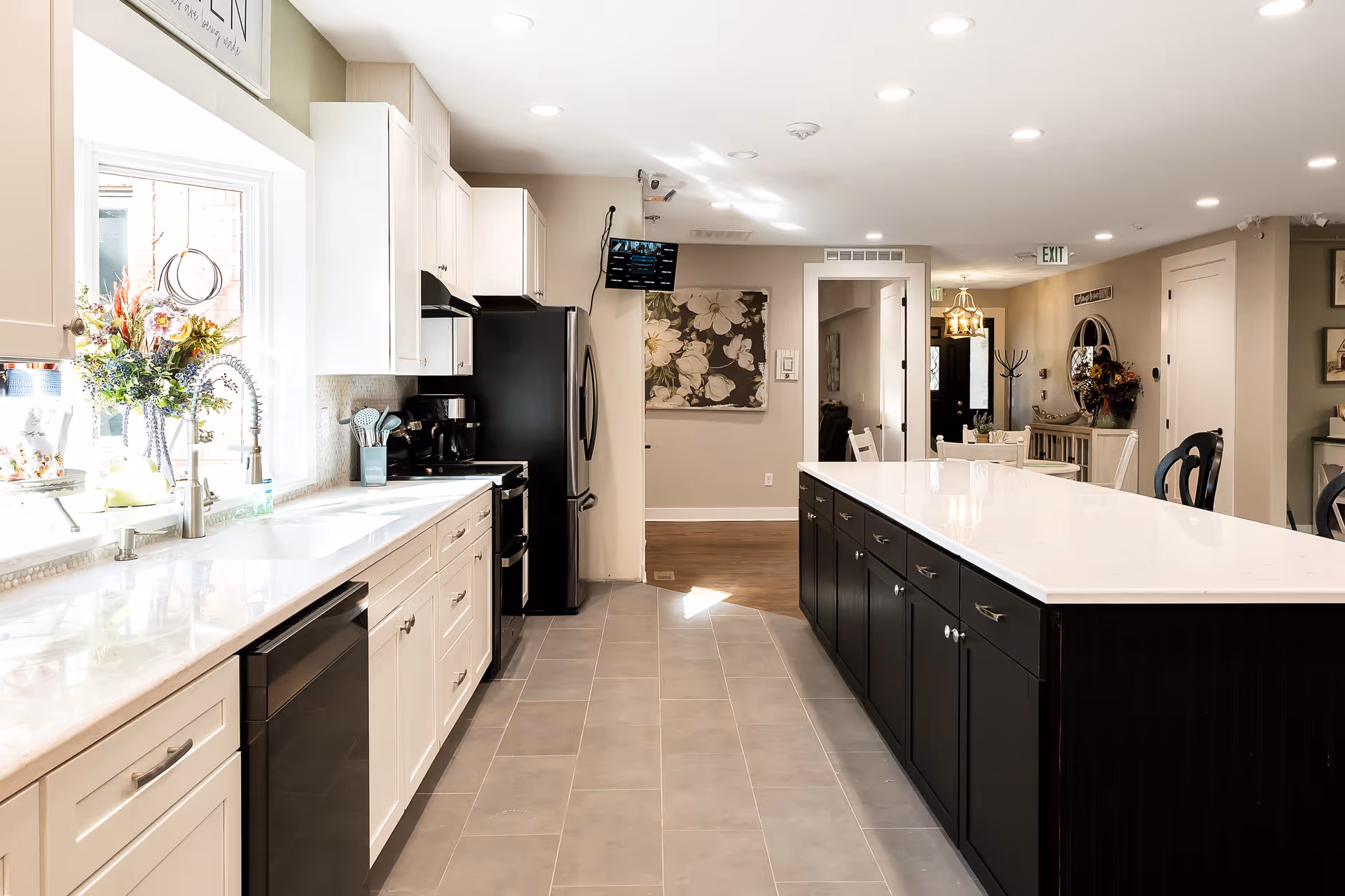 Bright and modern kitchen with white upper cabinets and black lower cabinets on a large island. The kitchen features a stainless steel refrigerator, stove, dishwasher, and a sink under a window with a floral arrangement. The floor is tiled, and the kitchen opens into a dining area with chairs and a table visible in the background.