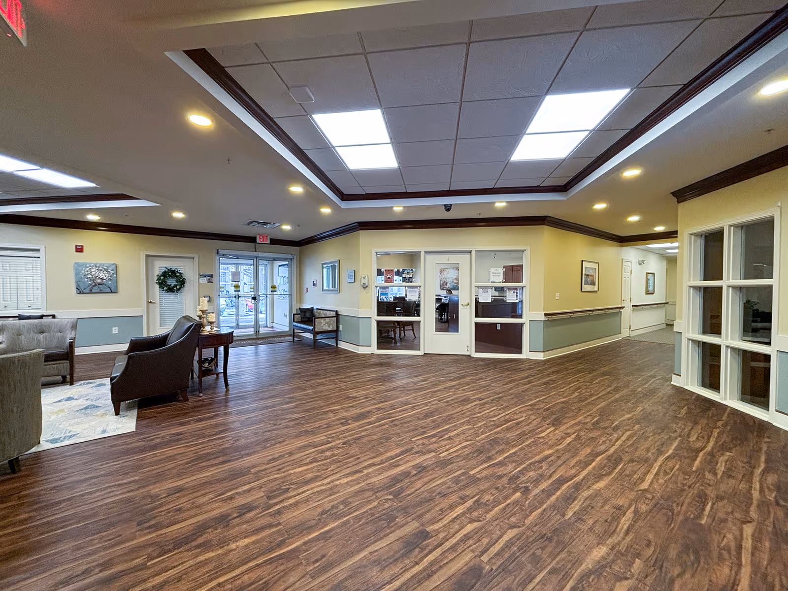 Spacious senior living facility lobby with wood flooring, beige and green walls, recessed lighting, and a large skylight. There are seating areas with chairs and a small table, glass double doors leading outside, and an office area with glass windows and doors.