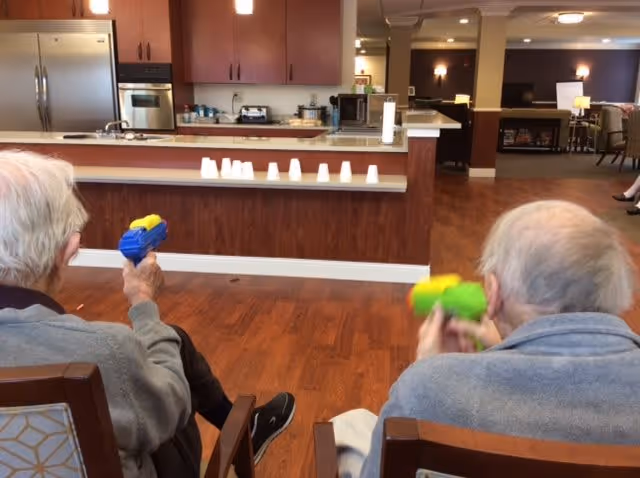 Two elderly individuals sitting on chairs indoors, each holding a small colorful toy gun and aiming at a row of white plastic cups lined up on a counter in front of them. The setting appears to be a common area with a kitchen in the background and a living room area visible further back.