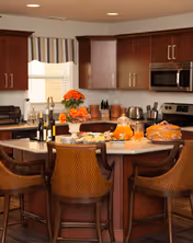 A modern kitchen with dark wood cabinets and stainless steel appliances including a microwave and stove. A kitchen island with a granite countertop is surrounded by four wicker bar stools. The island is decorated with a vase of orange flowers, bottles of wine, glasses, a pitcher of orange juice, and a cake covered with a glass dome. A window with a striped valance is above the sink.