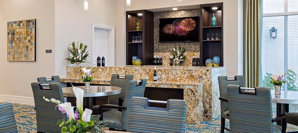 A well-lit interior lounge area with a granite countertop bar, decorated with vases of flowers and bottles. There are several striped upholstered chairs around round tables with floral centerpieces. A flat-screen TV displaying fireworks is mounted on the wall behind the bar, which is flanked by dark wooden shelves holding decorative items and glassware. Large windows with green curtains allow natural light into the space.