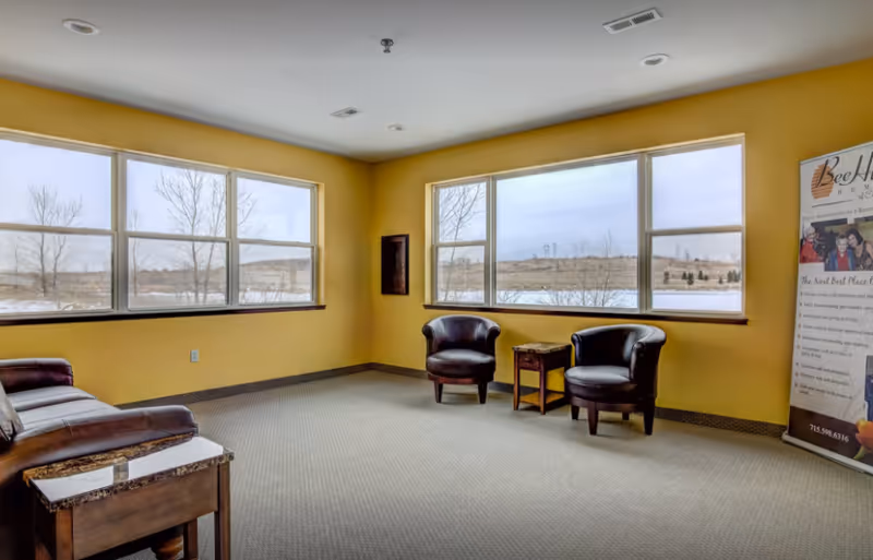 Bright common room with yellow walls, leather chairs, a small table, and large windows overlooking an outdoor landscape.