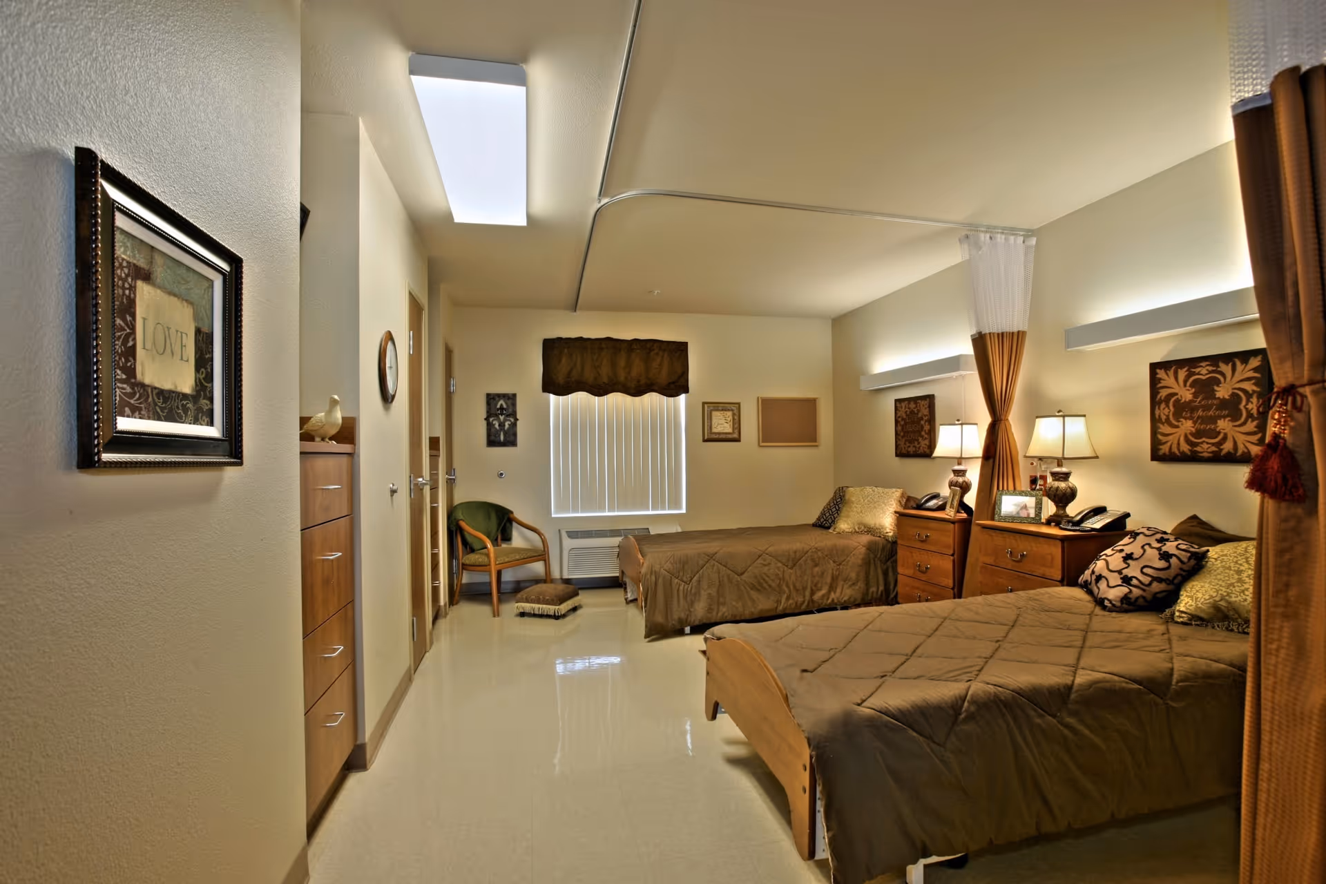 Shared bedroom with two twin beds, brown bedding, nightstands with lamps, a chair by the window, and wall art.
