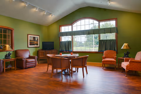 A cozy living room with green walls and a large window with green curtains. The room features a wooden floor, a round wooden table with four chairs, two orange upholstered armchairs, a brown leather armchair, a wooden TV stand with a television, two side tables with lamps, and a framed picture on the wall.