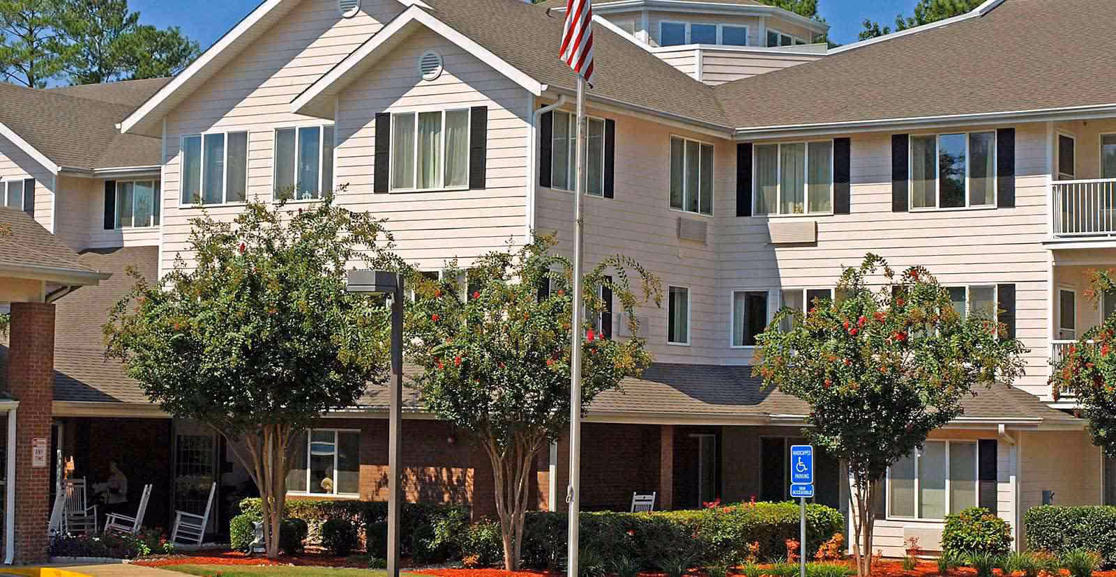 Exterior view of a multi-story senior living facility building with beige siding and brown roof. The building is surrounded by neatly trimmed bushes, small trees with red flowers, and a well-maintained garden area. There is an American flag on a flagpole in front of the building and a handicapped parking sign visible near the entrance.