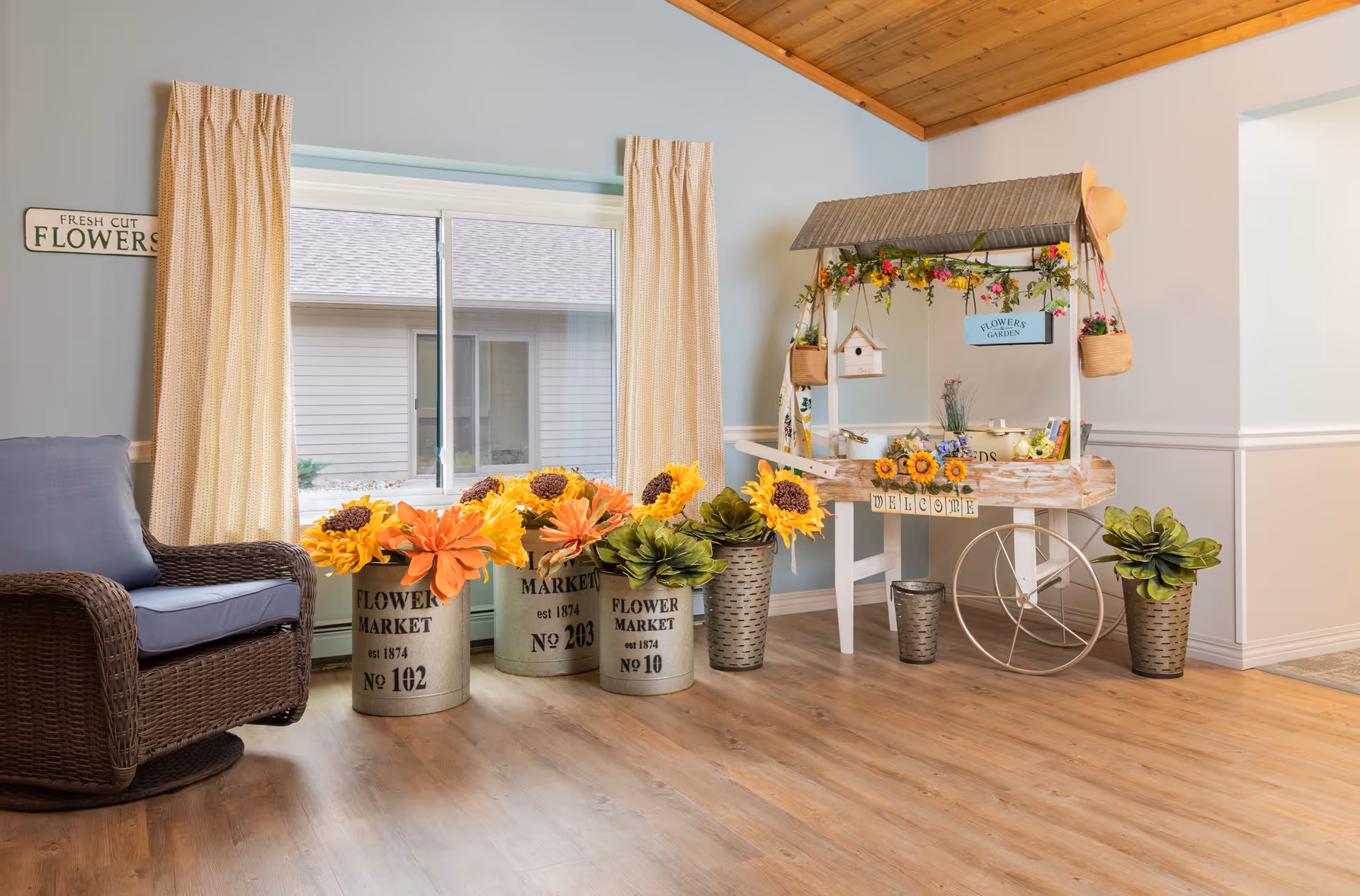 A cozy indoor corner with a wicker chair with a blue cushion, three metal flower pots labeled 'Flower Market' filled with large artificial sunflowers and orange flowers, and a decorative wooden flower cart adorned with hanging flower baskets, a birdhouse, and a 'Welcome' sign. The room has light blue walls, beige curtains, a wooden ceiling, and light wood flooring.