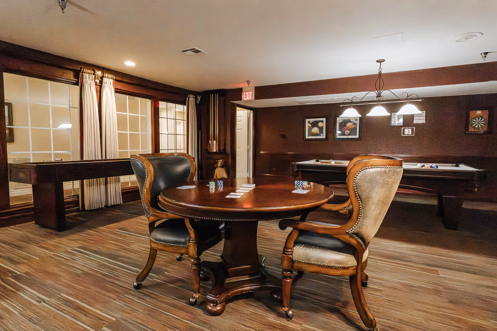 A cozy game room with a round wooden table and two upholstered chairs in the foreground. The table has playing cards and poker chips on it. In the background, there is a pool table with balls arranged on it and a shuffleboard table near large windows with curtains. The walls are decorated with framed pictures and a dartboard, and the room has warm lighting from ceiling fixtures.