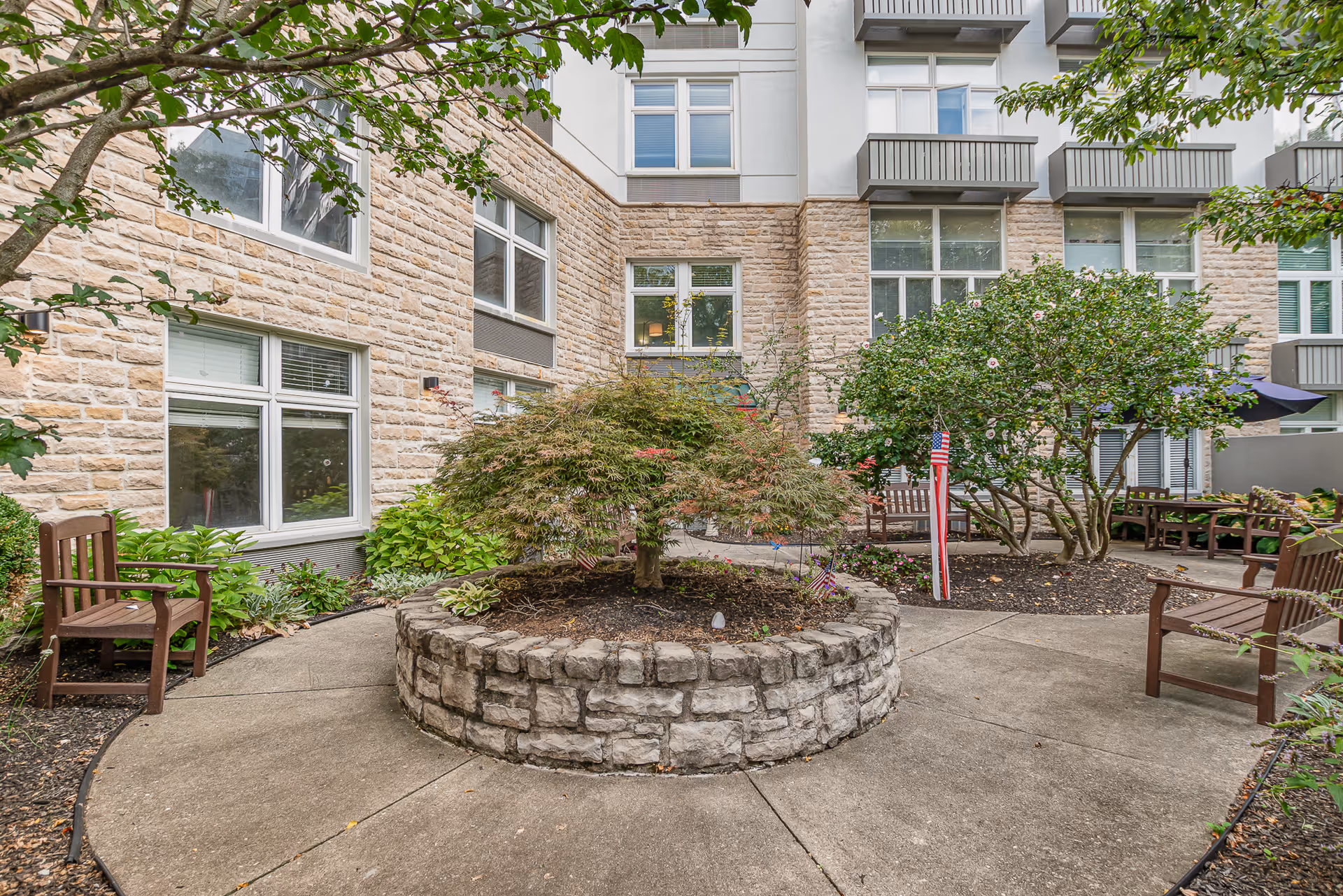 Courtyard at Wesley Glen with a circular stone planter, benches, small trees and shrubs, and apartment windows in the background.