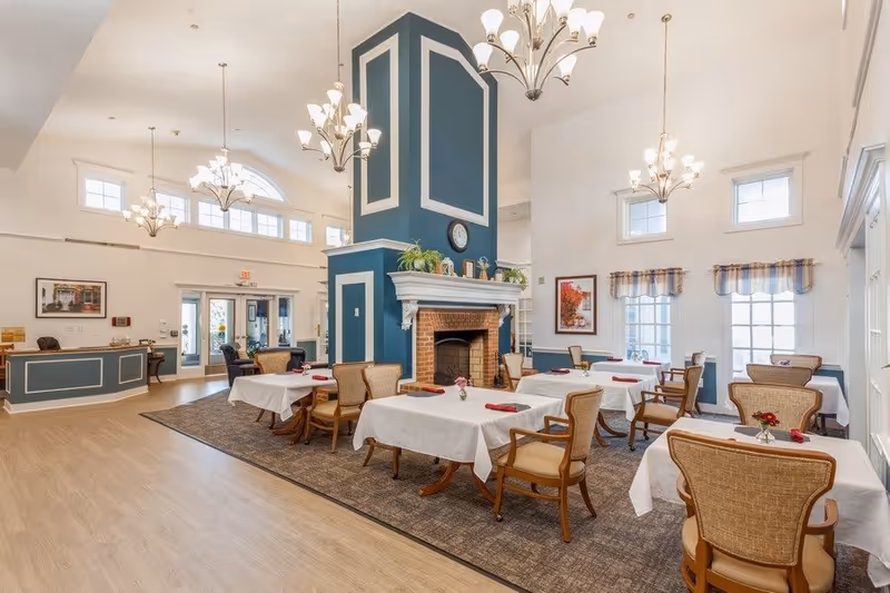 Bright dining room with tables set in white linens and chairs arranged around a central blue fireplace under high ceilings and chandeliers.