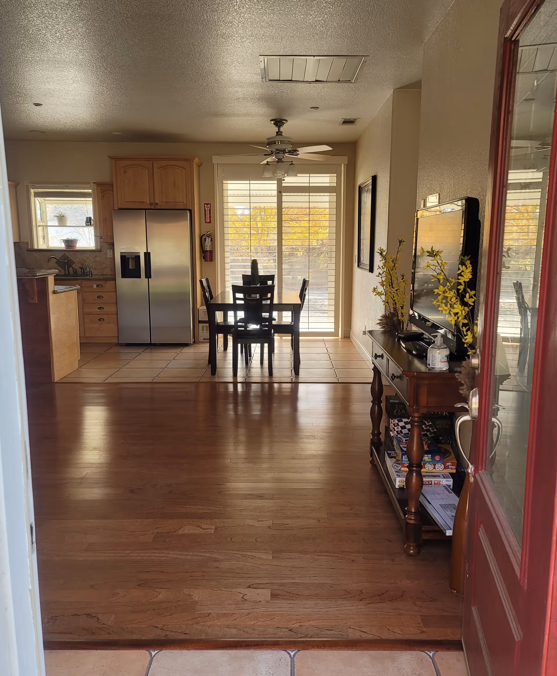View through an open red door into a home interior showing a wooden floor leading to a dining area with a table and four chairs. Beyond the dining area is a kitchen with wooden cabinets and a stainless steel refrigerator. A ceiling fan is above the dining table, and a sliding glass door with blinds lets in natural light. To the right, there is a wooden console table with a TV, decorative yellow flowers, and board games on the lower shelf.