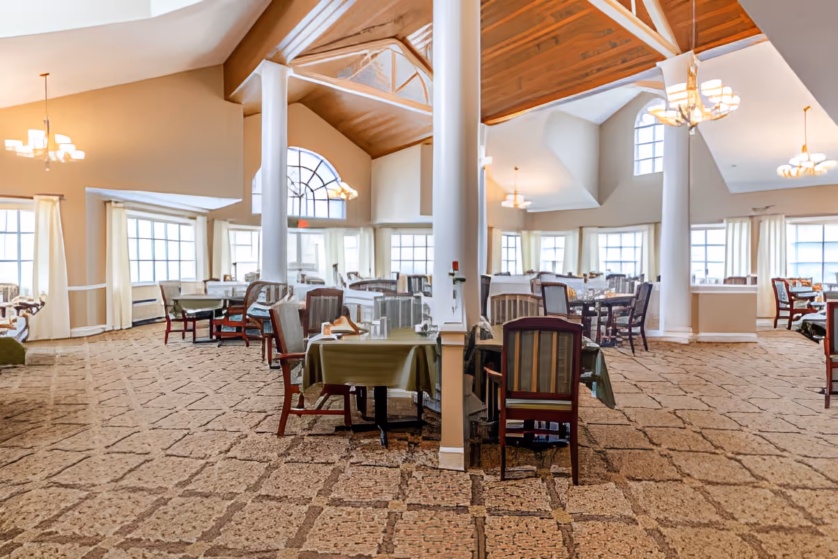 Spacious dining room with multiple tables and chairs arranged neatly. The room features large windows with cream-colored curtains allowing natural light to fill the space. The ceiling is vaulted with wooden accents and several chandeliers providing warm lighting. The carpet has a patterned design, and white columns add architectural interest to the room.