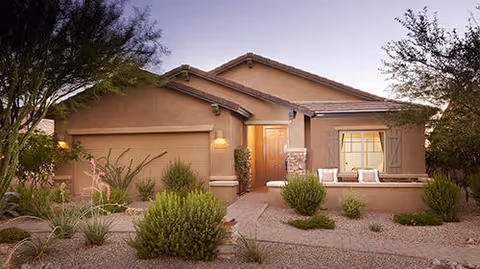 Exterior view of a single-story house with a two-car garage, a front door with a light above it, and a small seating area with cushions on the right side. The landscaping includes desert plants and gravel, with trees framing the house under a clear evening sky.