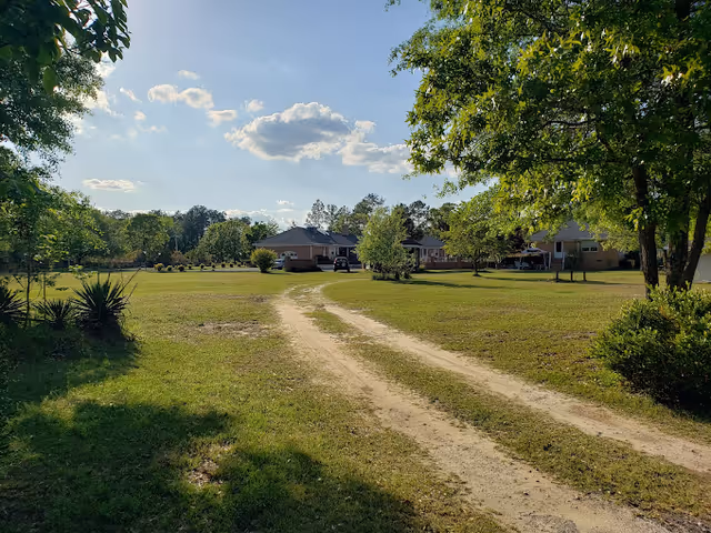 A dirt pathway leads through a grassy area with trees on both sides toward a group of single-story buildings under a partly cloudy blue sky.