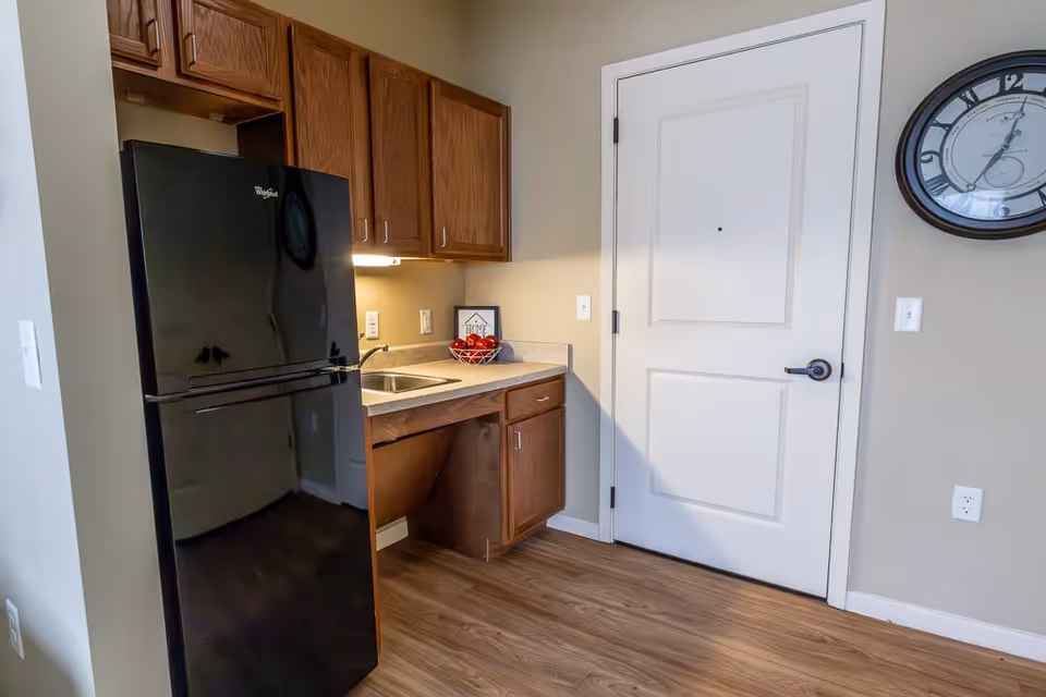 A small kitchen area with a black Whirlpool refrigerator, wooden cabinets above and below a countertop with a sink. There is a bowl of red apples and a framed picture on the counter. A white door with a black handle is next to the kitchen area, and a round wall clock is mounted on the beige wall above a light switch and electrical outlet. The floor is wood laminate.