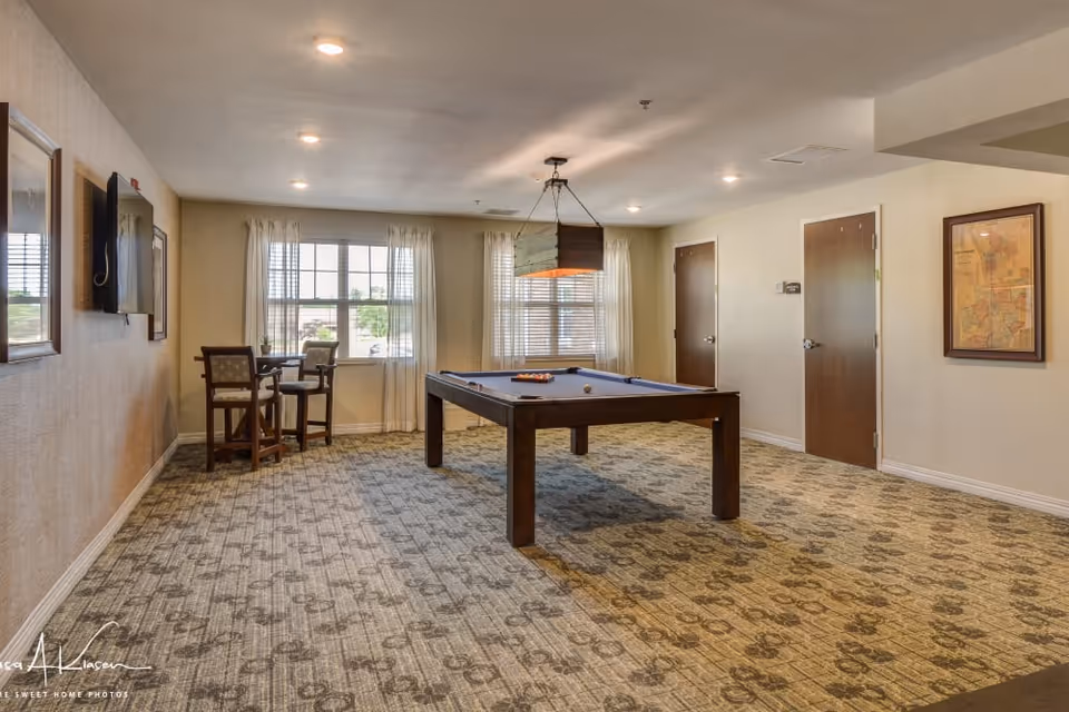 A spacious room with a pool table in the center, a hanging light fixture above it, a small table with two chairs near the window, beige walls, patterned carpet, and two closed wooden doors on the right wall.