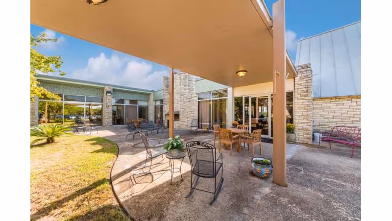 Outdoor patio area at Hilltop Village Nursing and Rehab with metal and wooden chairs and tables, a covered section with ceiling lights, potted plants, and a stone building exterior under a clear blue sky.