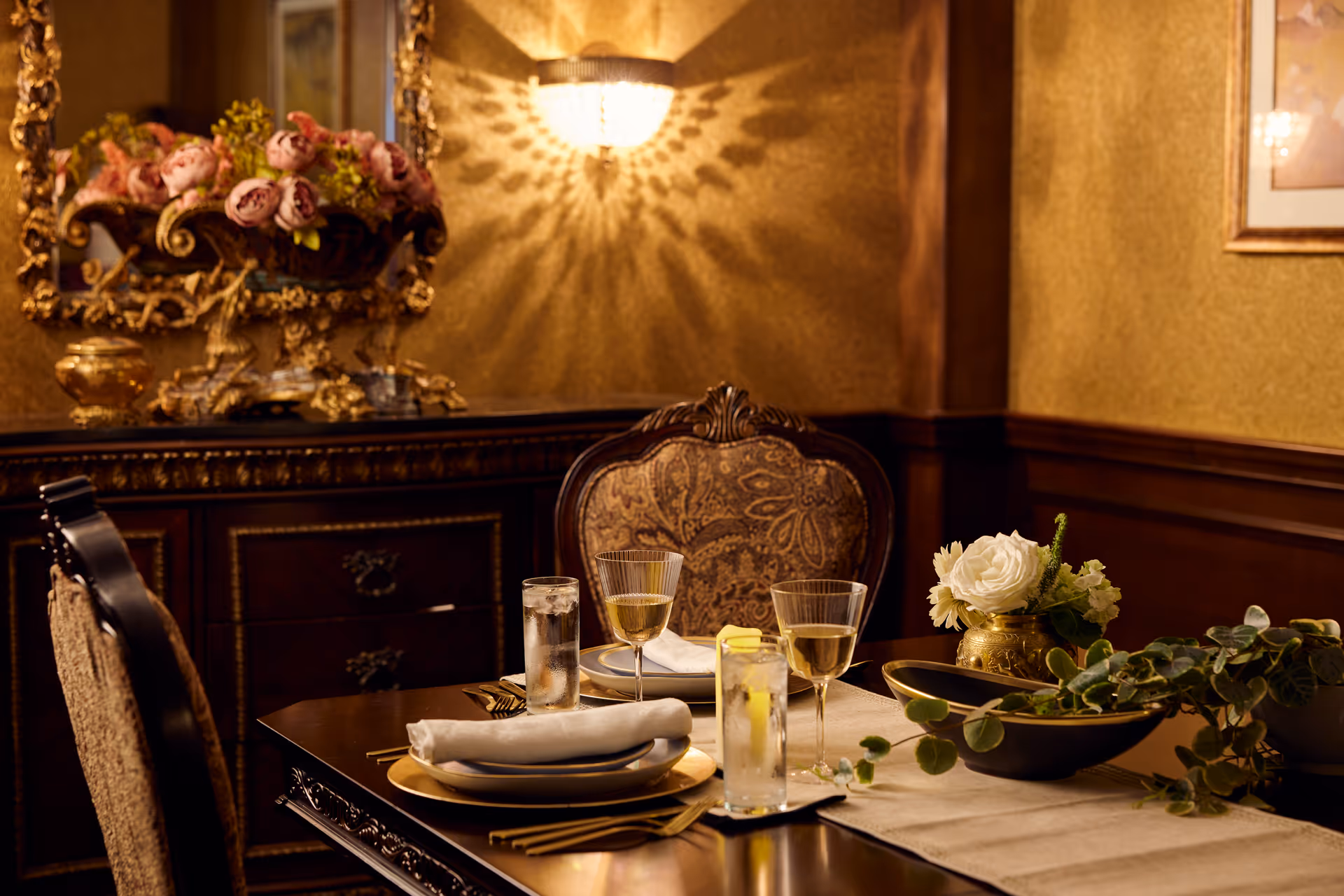 A warmly lit dining room with an elegant wooden table set for two, featuring gold-rimmed plates, folded white napkins, glasses of water with lemon, and wine glasses. The background includes a decorative sideboard with a large ornate gold-framed mirror and a floral arrangement. The walls are adorned with a golden textured finish and a wall sconce casting a patterned light.