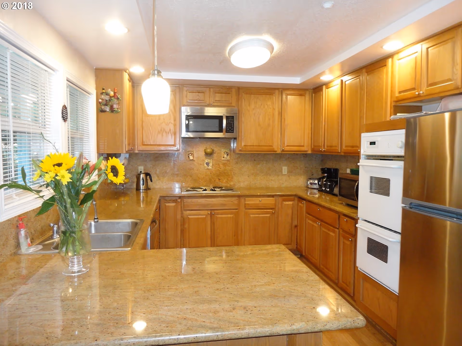 A well-lit kitchen with wooden cabinets and granite countertops. The kitchen features a double sink, a microwave above a stovetop, a double oven, a stainless steel refrigerator, and small appliances like a coffee maker and toaster. A vase with sunflowers is placed on the countertop near the sink. There are windows with blinds on the left side and ceiling lights providing illumination.