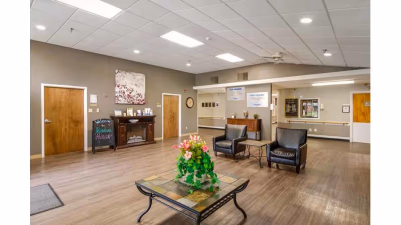 A spacious and well-lit common area in a senior living facility with wooden floors, beige walls, and a white ceiling with recessed lighting. The room features a small table with a flower arrangement in the center, two black leather chairs with a small side table between them, a wooden cabinet with decorative items, and a chalkboard sign that says 'Welcome Broadview Health and Rehabilitation'. There are several wooden doors and informational posters on the walls.