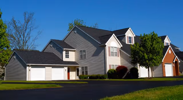 Two-story townhouse-style senior living building with attached garages, driveway, lawn, and a clear blue sky.