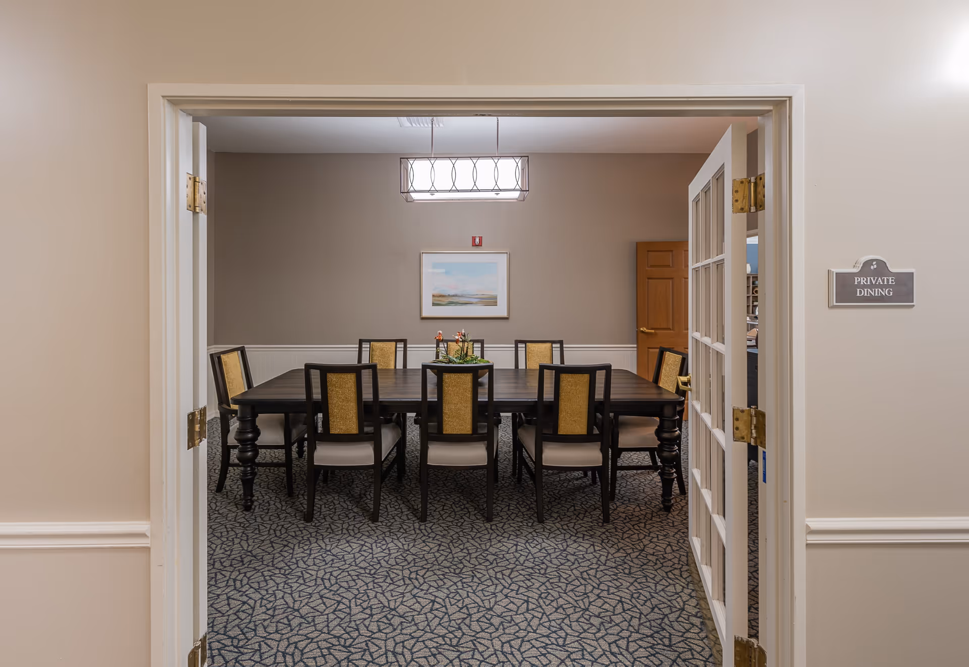 View through open double doors into a private dining room with a large rectangular wooden table surrounded by eight chairs. The room has beige walls, a patterned carpet, a framed landscape painting on the wall, and a modern rectangular ceiling light fixture.