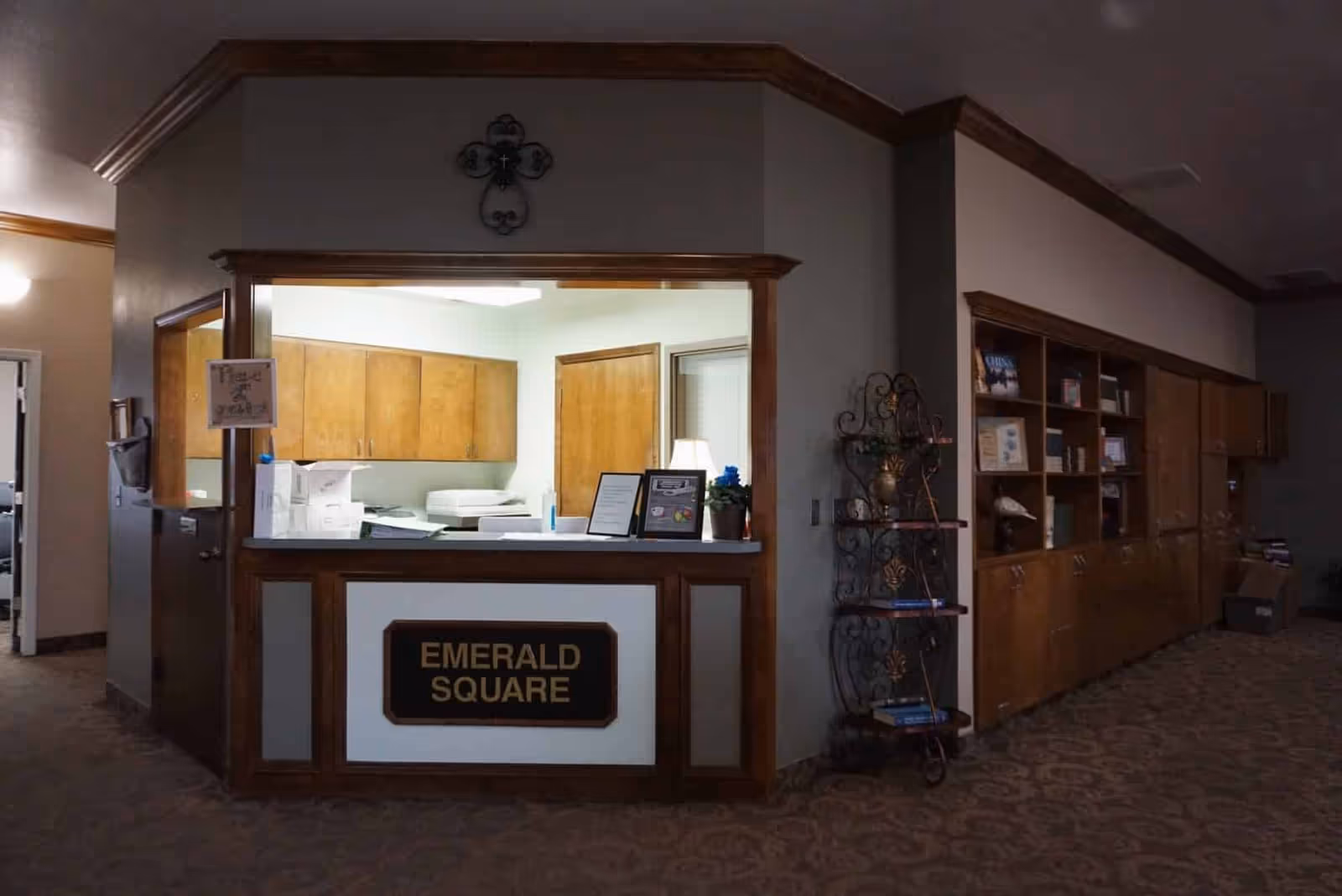 Reception area labeled Emerald Square with a counter and an open window into an office with wooden cabinets and a door. To the right, there is a decorative metal shelf with books and a long wooden cabinet with shelves and various items. The area is carpeted and has beige walls with wooden trim.