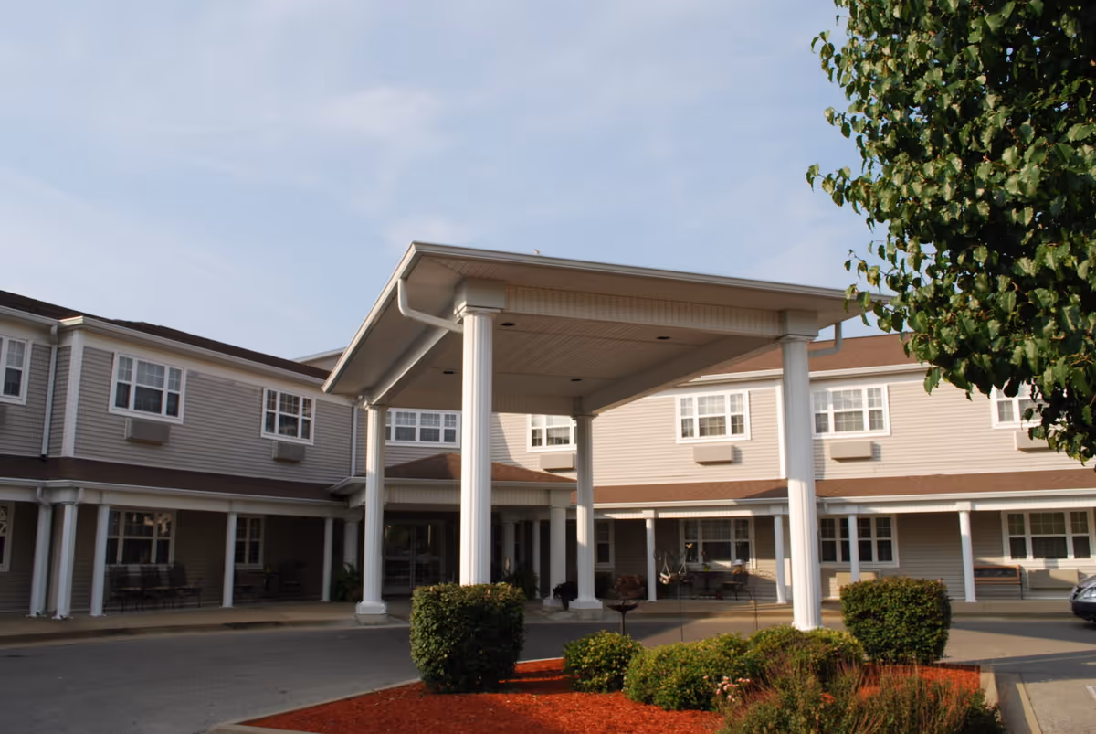 Entrance of a two-story senior living building with a covered porte-cochère, white columns, and landscaped beds in front.