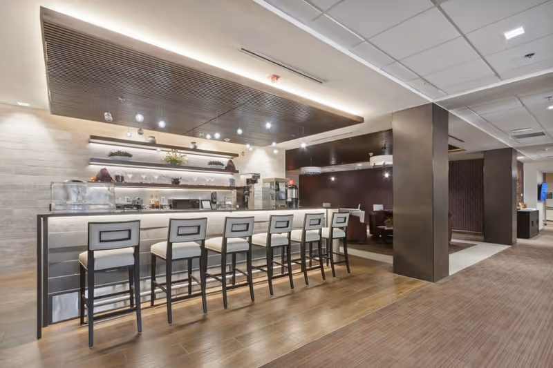 Modern communal bar and seating area with a row of high stools at a backlit counter in a senior living facility interior.