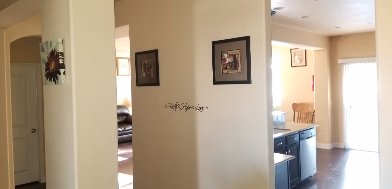 Interior view of a senior living facility showing a hallway with beige walls and framed pictures. To the left, there is a glimpse of a living room with a dark brown leather sofa. To the right, there is a kitchen area with dark cabinets, a dishwasher, and a wooden chair near a sliding glass door letting in natural light. The wall in the center has a decorative text that reads 'Faith Hope Love'.