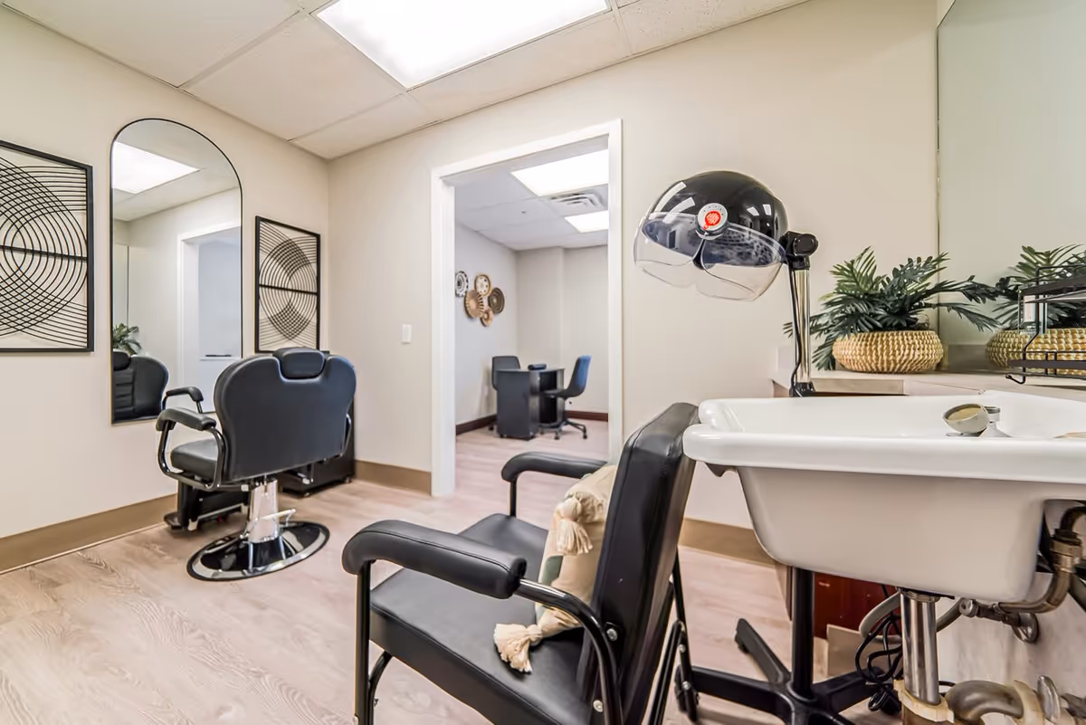 A salon-style interior with barber chairs, a hooded hair dryer, sink basin, wall mirrors, and decorative plants.