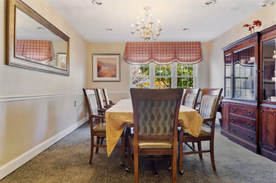 A dining room with a rectangular table covered by a yellow tablecloth surrounded by eight wooden chairs with patterned upholstery. A large mirror hangs on the left wall, and a framed picture is on the far wall. A window with red and white checkered valance lets in natural light. On the right side, there is a wooden china cabinet with glass doors displaying dishes. A chandelier hangs from the ceiling.