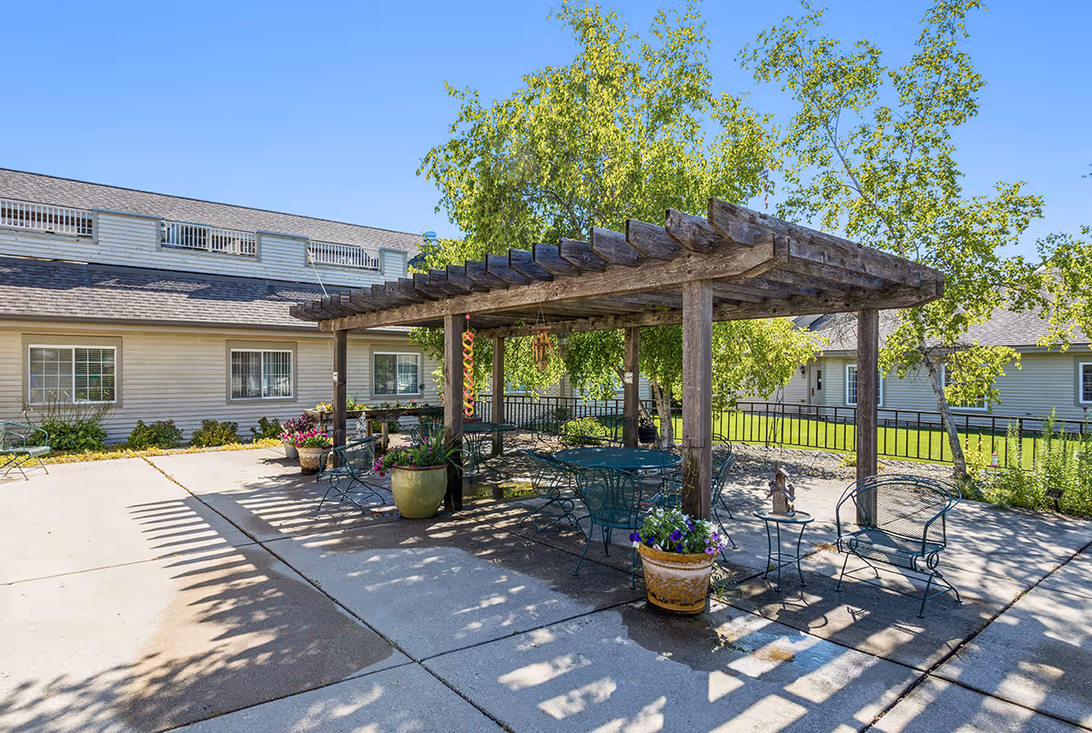 Outdoor patio area with a wooden pergola providing shade over metal tables and chairs. There are potted plants with flowers around the seating area, and trees with green leaves in the background. The patio is adjacent to a light-colored building with multiple windows under a clear blue sky.