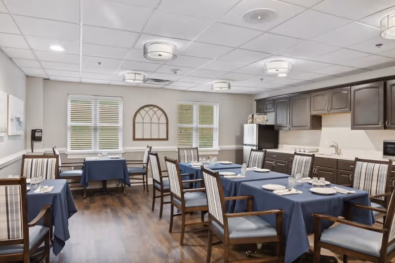 A dining room with several tables covered in blue tablecloths, each set with plates, glasses, and napkins. The room has wooden chairs with striped cushions, two windows with blinds, and dark wood cabinets along one wall with a white refrigerator and microwave. The ceiling has multiple round light fixtures.
