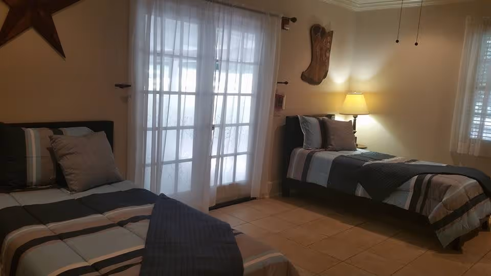 Two twin beds in a softly lit bedroom with a lamp, sheer-curtained French doors, and a tiled floor.