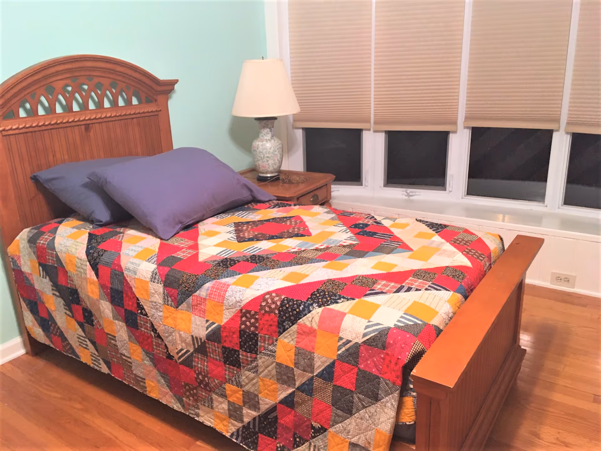 Wood-framed bed with a colorful patchwork quilt and purple pillows beside a nightstand and lamp in a room with windows and blinds.