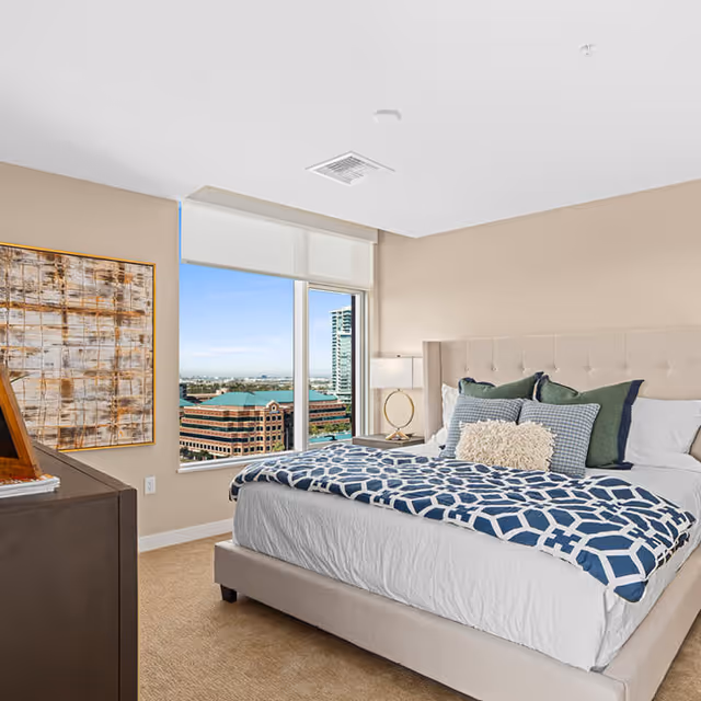 A bright bedroom with a large bed featuring a beige upholstered headboard, white bedding, and a blue patterned throw blanket. Several decorative pillows are arranged on the bed. A nightstand with a modern lamp is next to the bed. A large window offers a view of nearby buildings and a clear blue sky. A piece of abstract artwork hangs on the wall beside the window.