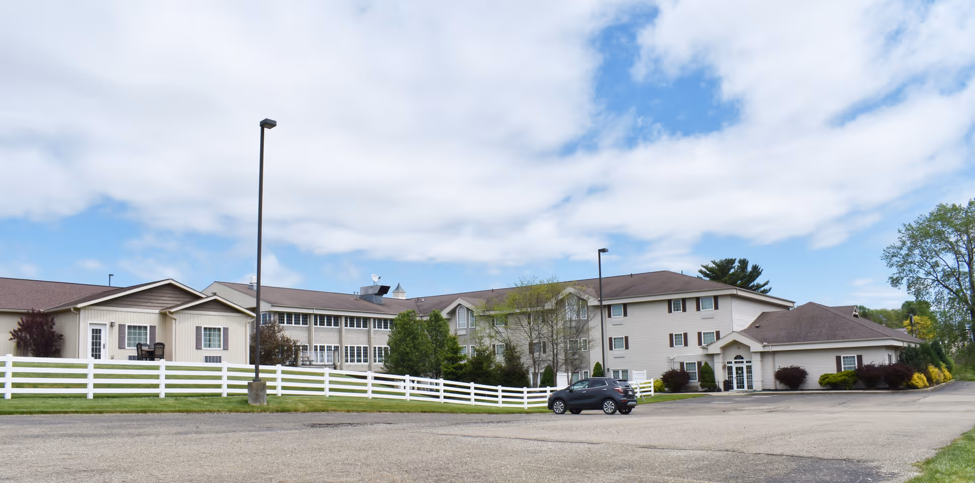 Exterior view of a senior living facility named Senior Suites at Century Farms, showing a large multi-story building with beige siding, multiple windows, and a white fence in front. There is a parking lot with a single parked car and a partly cloudy sky overhead.