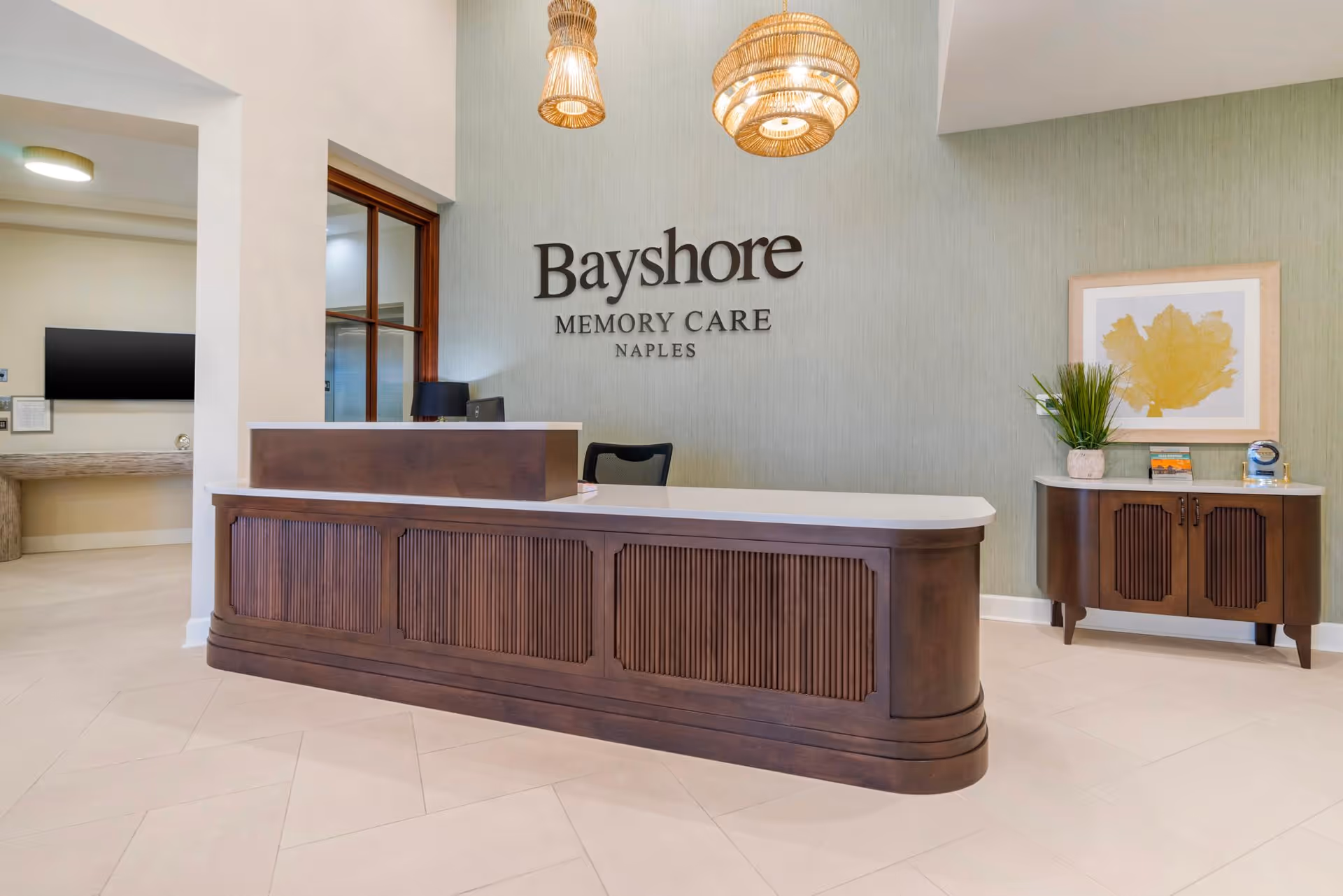 Reception area of Bayshore Memory Care in Naples featuring a wooden front desk with a white countertop, a chair behind the desk, two hanging woven pendant lights, a side cabinet with a plant and framed artwork, and the facility name displayed on a light green textured wall.