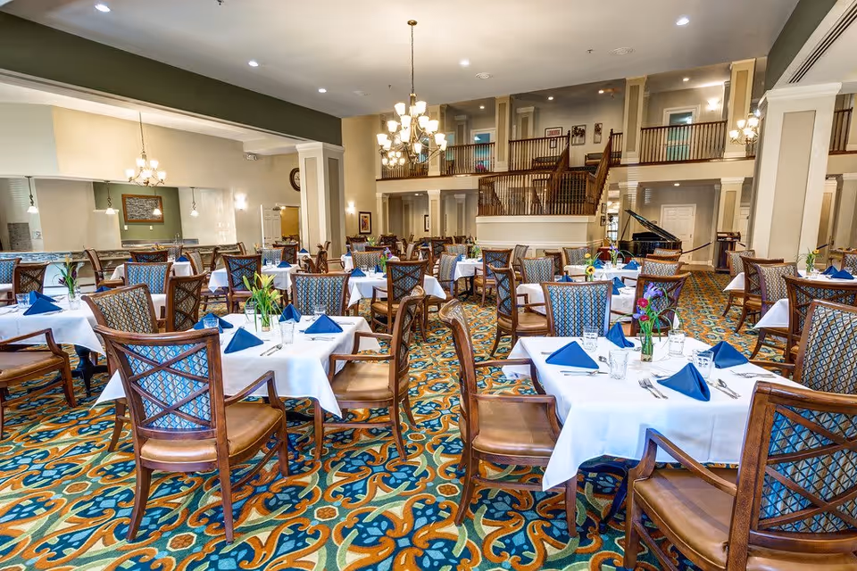 A spacious dining room with multiple tables covered in white tablecloths, each set with blue folded napkins, glasses, and silverware. The room features patterned carpet, wooden chairs with cushioned seats, chandeliers hanging from the ceiling, and a grand piano in the background near a staircase leading to an upper level.