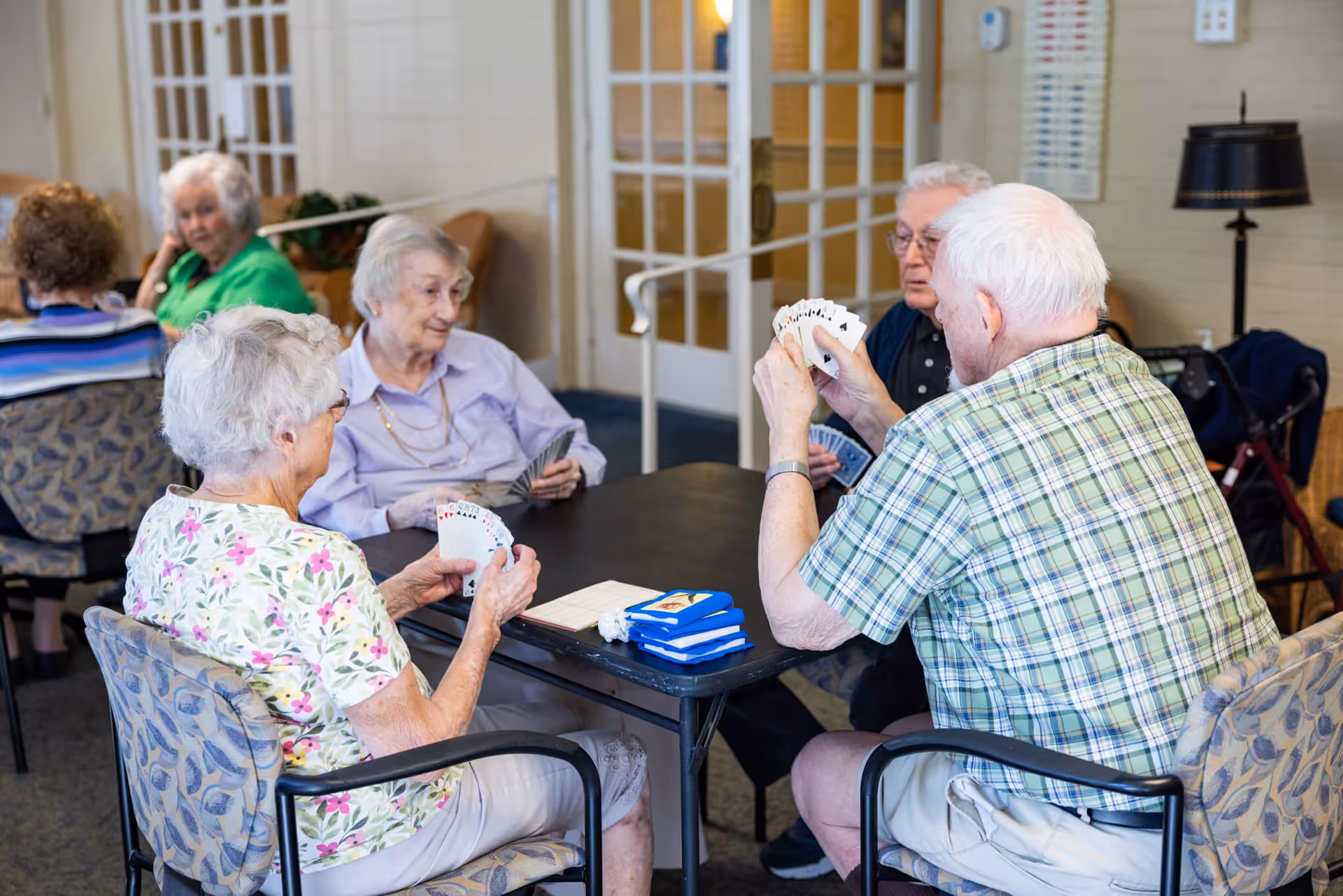 Four elderly people sitting around a table playing cards in a well-lit room with patterned chairs and glass-paneled doors in the background. Two women and two men are engaged in the game, with additional elderly individuals visible in the background.
