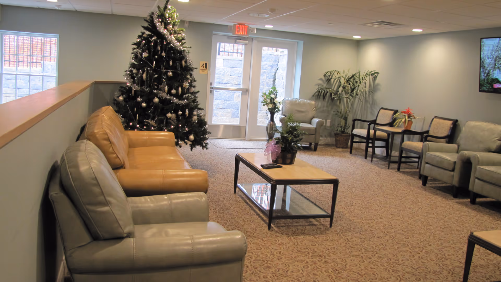 A cozy living room area decorated for the holidays with a Christmas tree adorned with silver ornaments and garlands. The room features several armchairs and a sofa arranged around a wooden coffee table with potted plants. There are large windows and a glass door letting in natural light, and a flat-screen TV mounted on the wall.