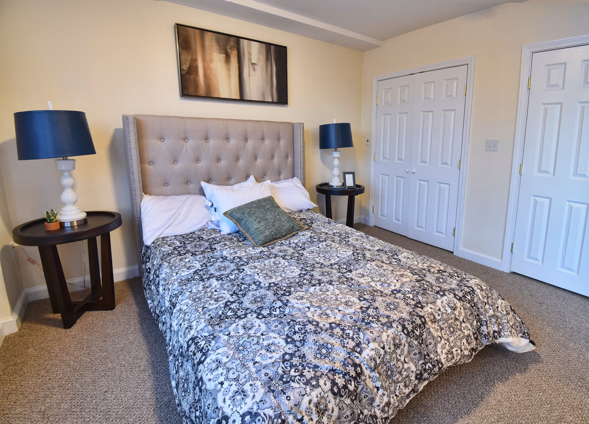 A bedroom with a large bed featuring a tufted headboard, patterned bedspread, and multiple pillows. There are two round bedside tables, each with a white lamp with a dark blue shade. The room has beige walls, carpeted floor, and two white double-door closets.