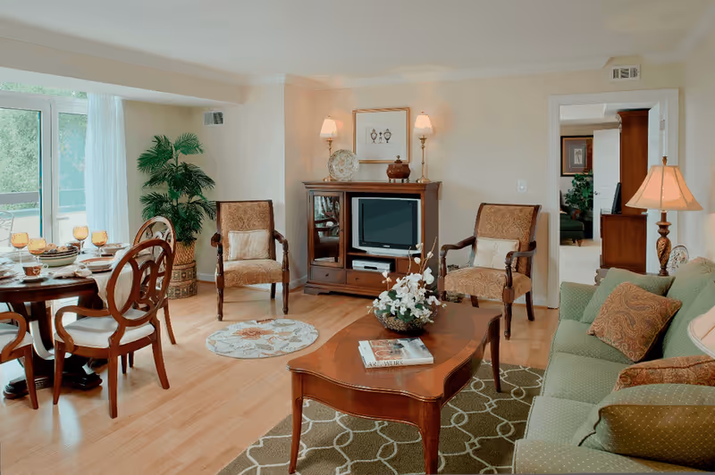 A bright living room and dining area with a green sofa, wooden coffee table, TV cabinet, and a dining set by a large window.