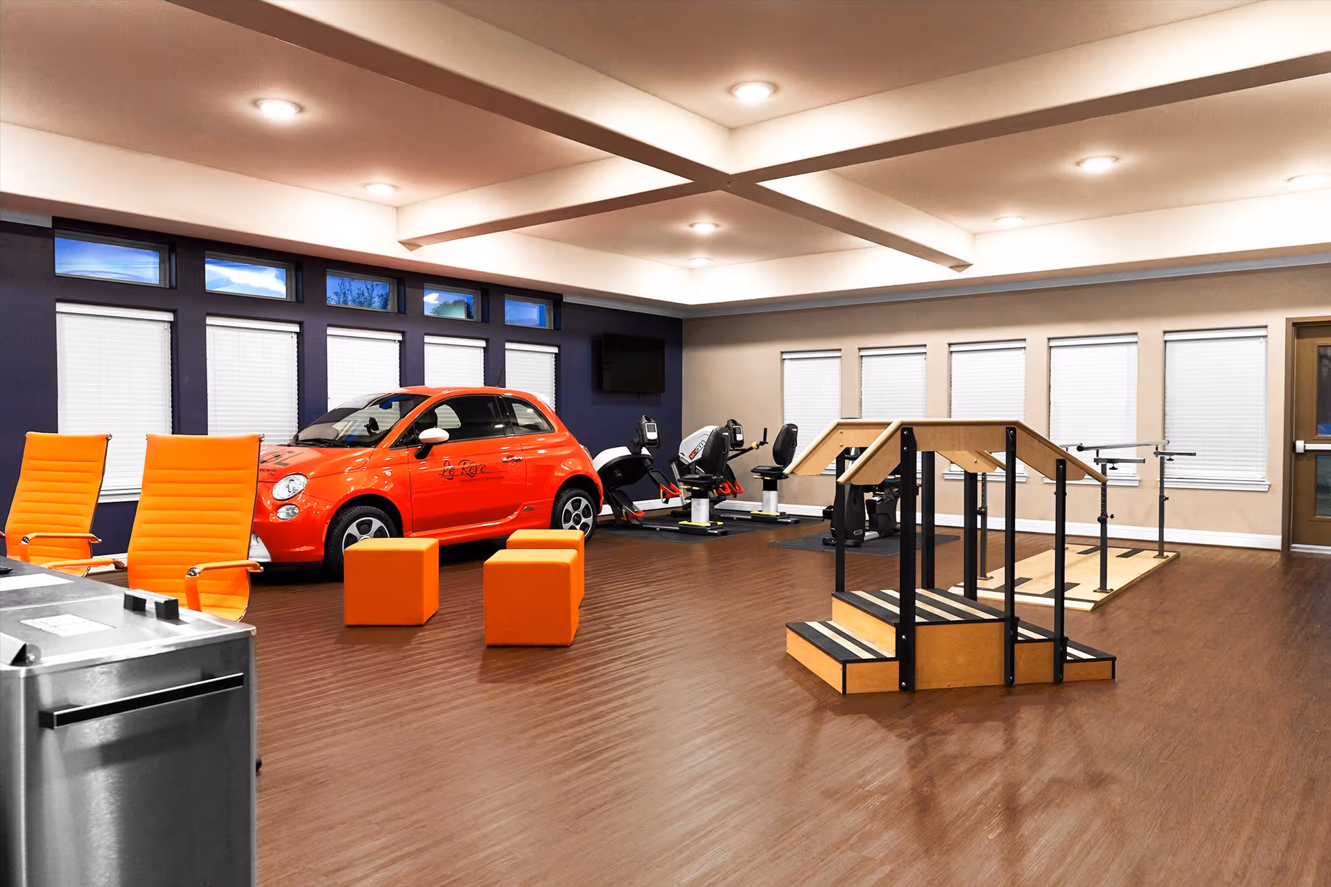 A spacious rehabilitation room with wooden flooring featuring a bright orange compact car, two orange chairs, two orange cube seats, exercise equipment including stationary bikes, and a wooden staircase with handrails for physical therapy. The room has multiple windows with blinds and recessed ceiling lights.
