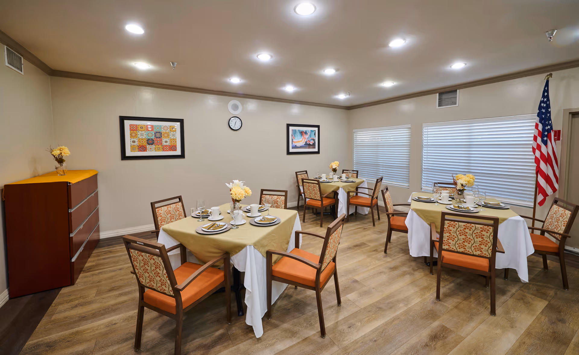 A dining room with four square tables covered with beige and white tablecloths, each set with plates, cups, and silverware. The room has wooden chairs with orange cushions and patterned backs, light wood flooring, two framed pictures on the wall, a clock, and an American flag in the corner near two windows with closed blinds.