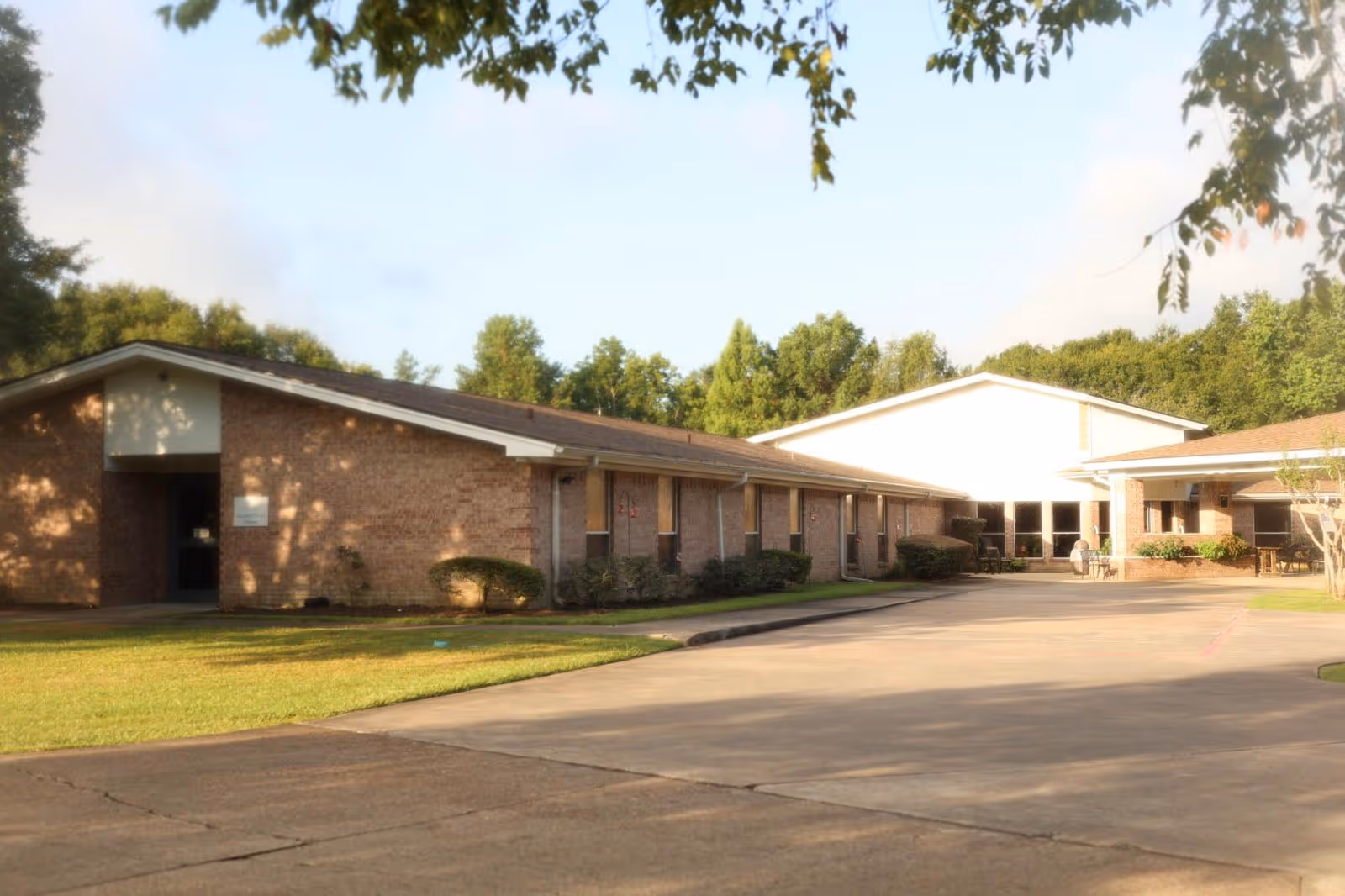 Exterior view of a single-story brick building with a sloped roof, surrounded by green grass and trees under a clear sky. The building appears to be part of a senior living facility with a paved driveway in front.