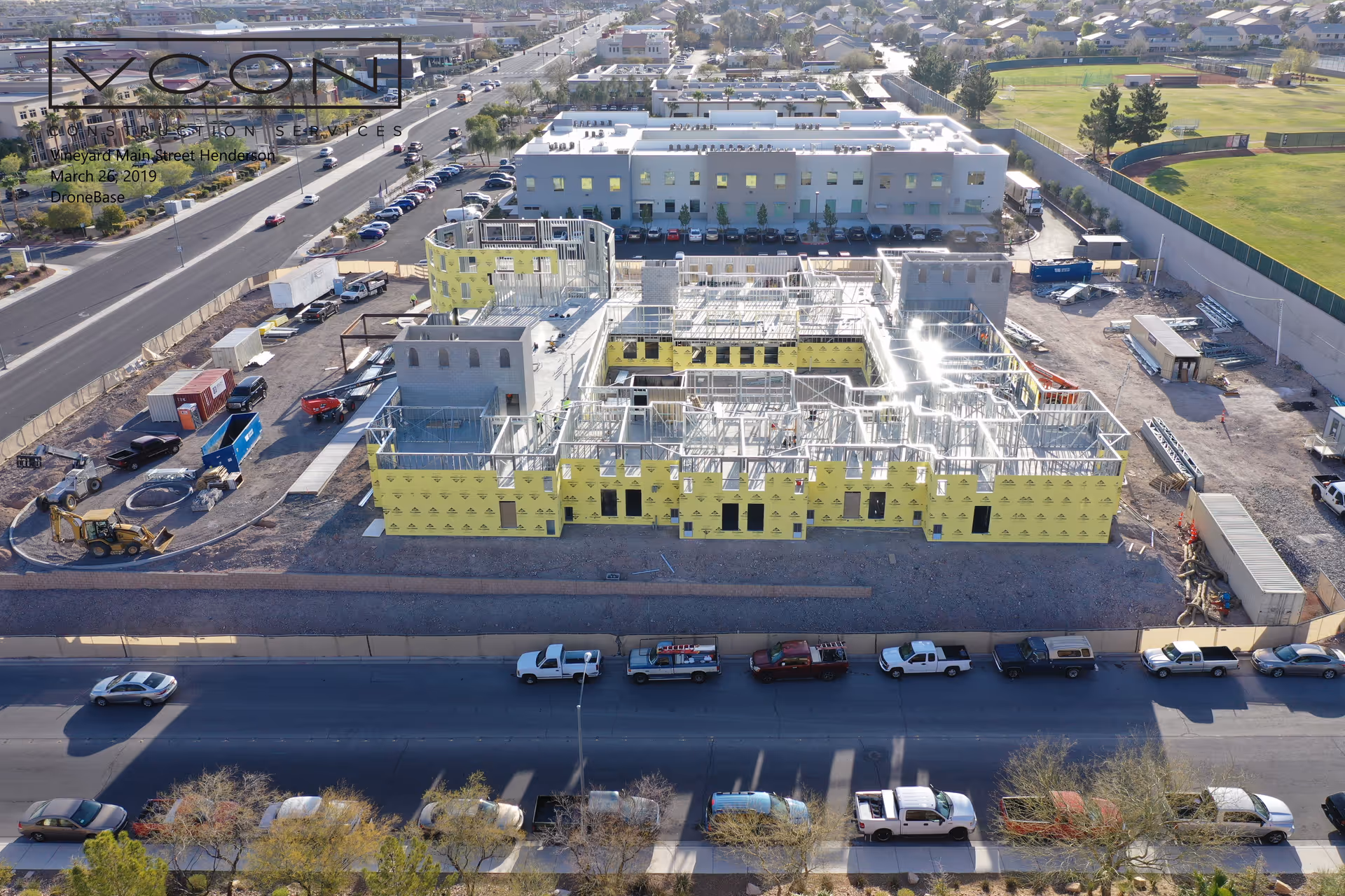 Aerial view of a construction site for a building with yellow exterior insulation panels and steel framing, surrounded by parked vehicles and adjacent to a road. In the background, there is a completed building, residential houses, and a green field with a baseball diamond.
