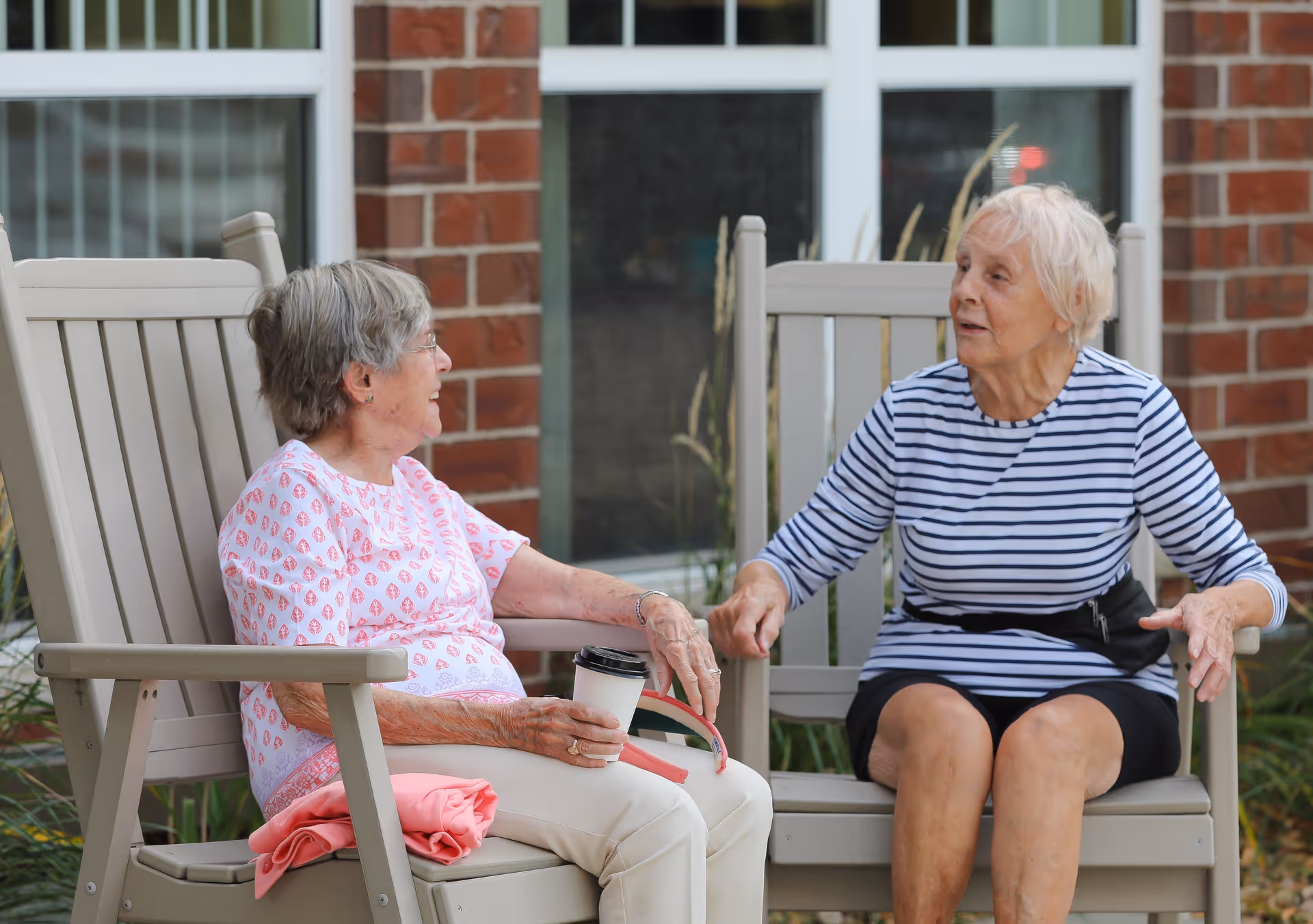 Two elderly women sitting outdoors on beige wooden chairs, engaged in conversation. One woman is wearing a white shirt with pink patterns and holding a coffee cup, while the other is wearing a striped long-sleeve shirt and black skirt. They are seated in front of a brick wall with windows.
